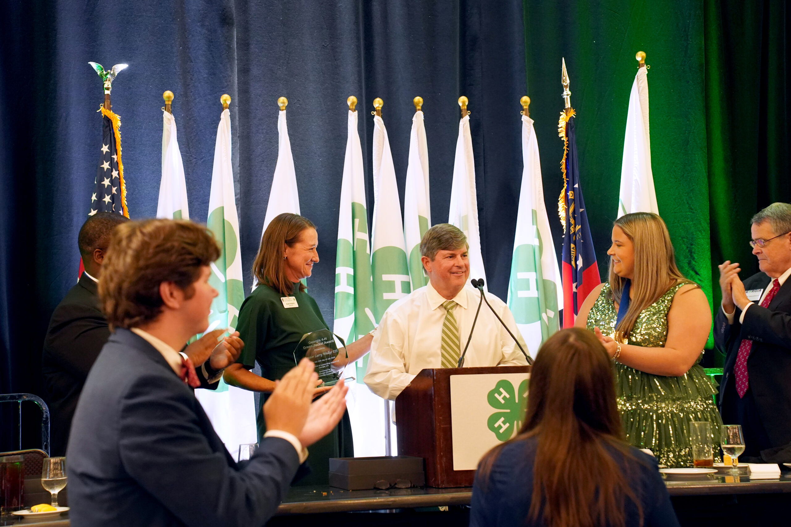 Larry Walker stands at a podium with 4-H flags behind him as he receives the Green Jacket Award. Others around him applaud and smile.