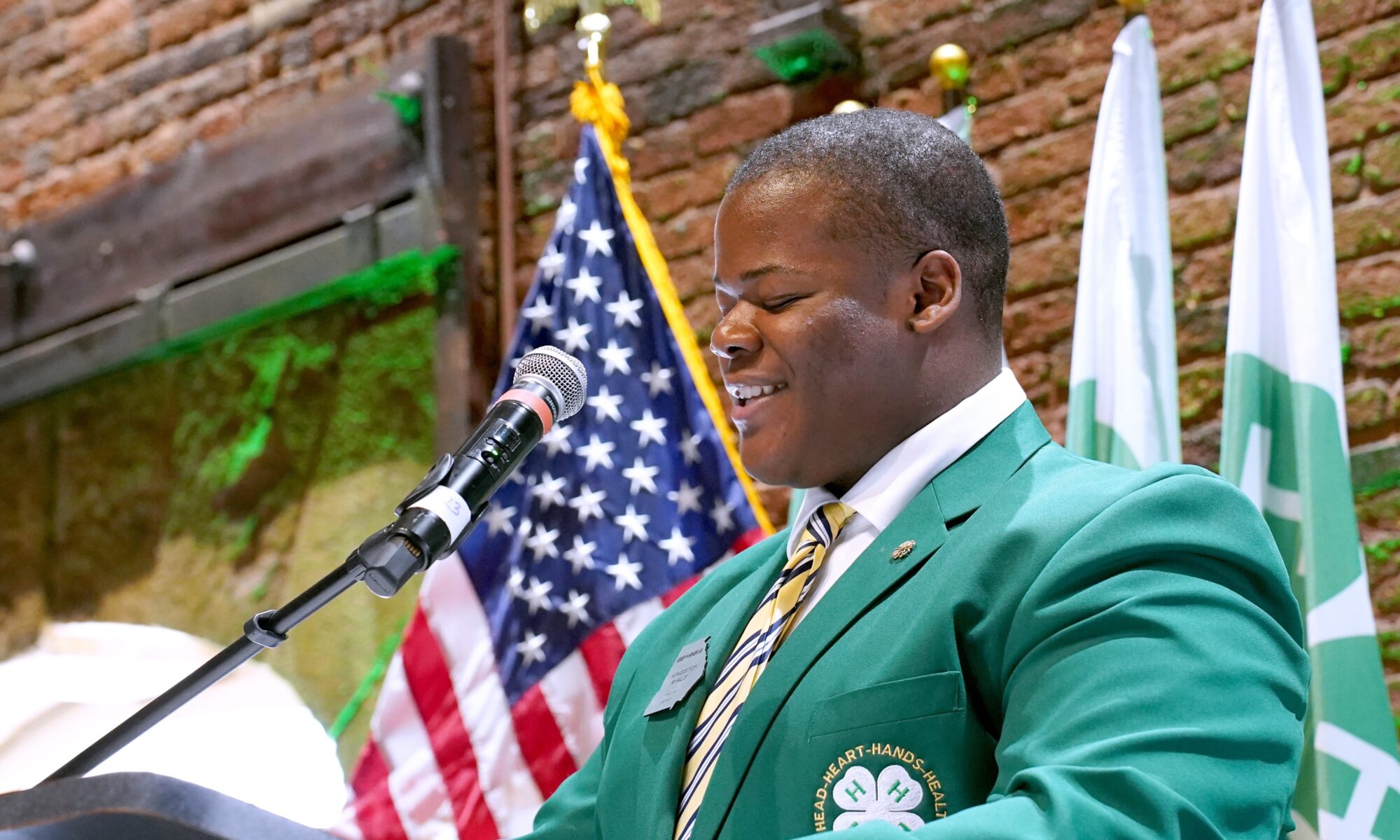 A photo of a student speaking at a podium in front of the American flag. The student speaking is wearing a green jacket and is speaking at the Georgia Railroad Freight Depot during Georgia 4-H Day at the Capitol.