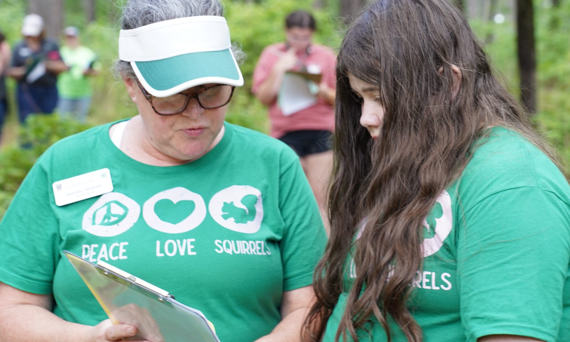 A photo of a student and an adult leader wearing green shirts looking at a clipboard in the woods. This photo was taken during the State Wildlife Judging competiton at Rock Eagle 4-H Center.