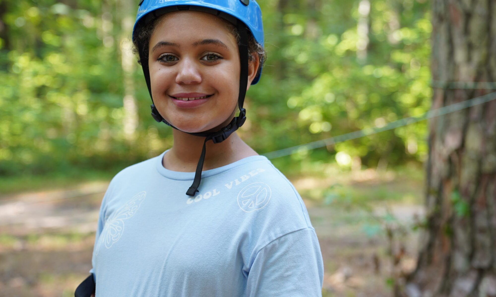 A photo of a student wearing a blue shirt and a blue helmet smiling while in the woods.